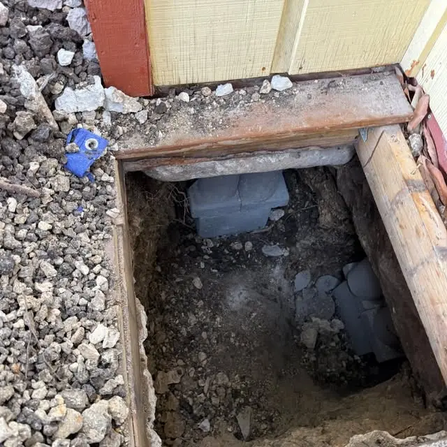 Excavated foundation access point showing concrete support blocks beneath a residential structure. The exposed area allows crews to inspect and stabilize foundation settlement.