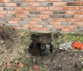 Foundation repair in progress with an excavated access point exposing concrete piers beneath a brick home. The opening provides access for foundation stabilization work.